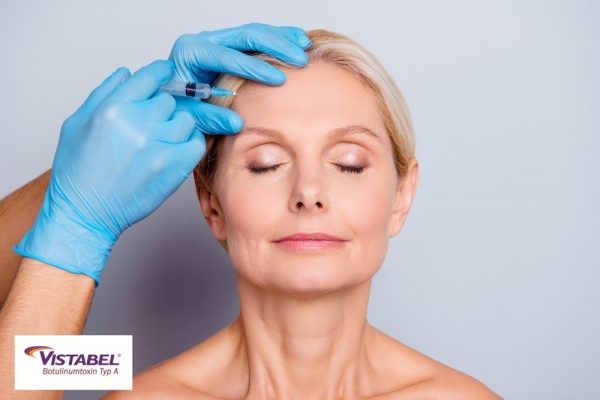Portrait of calm serious aged woman with wrinkle keeping eyes close getting injection in forehead in professional clinic isolated on grey background